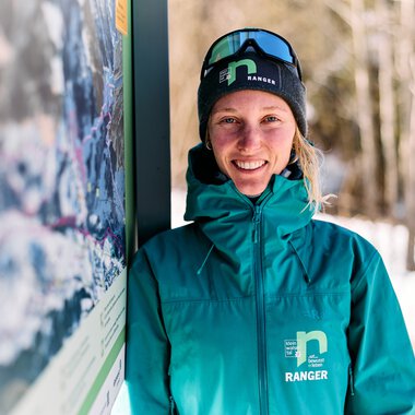Eine lächelnde Frau in Winterkleidung und mit Skibrille steht neben einem Informationsschild im Schnee, im Hintergrund Bäume und verschneite Landschaft. | © Kleinwalsertal Tourismus eGen • Fotograf: Oliver Farys