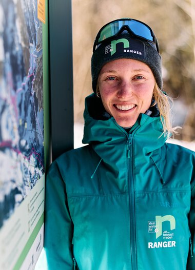 Eine lächelnde Frau in Winterkleidung und mit Skibrille steht neben einem Informationsschild im Schnee, im Hintergrund Bäume und verschneite Landschaft. | © Kleinwalsertal Tourismus eGen • Fotograf: Oliver Farys