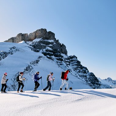 Five people snowshoeing across a snowy mountain landscape in Kleinwalsertal, with a high rock massif in the background and clear blue sky. | © Kleinwalsertal Tourismus eGen • Photographer: Oliver Farys