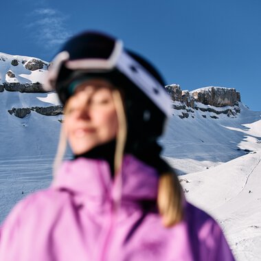 Blurred person wearing a ski helmet in winter with snowy mountains, rocks, and blue sky in the background. | © Kleinwalsertal Tourismus eGen • Photographer: Oliver Farys