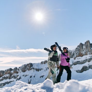 Two skiers standing on snowy mountains under a bright sun with clear blue sky and snow-covered peaks in the background. | © Kleinwalsertal Tourismus eGen • Photographer: Oliver Farys