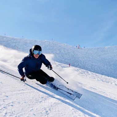 Skier in blue jacket and dark pants maneuvering during a turn on a snowy slope on a sunny winter day. | © Kleinwalsertal Tourismus eGen • Photographer: Oliver Farys