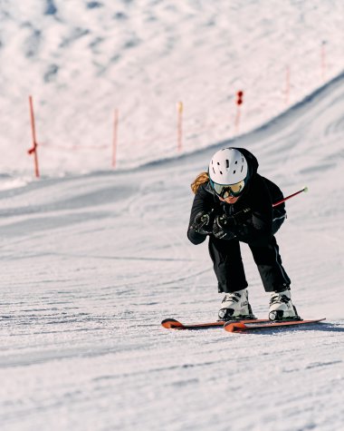 Ein Kind beim Skifahren auf einer Piste, tragen Helm und Skibrille, in winterlicher Schneelandschaft. | © Kleinwalsertal Tourismus eGen • Fotograf: Oliver Farys