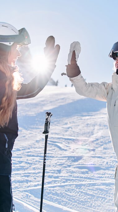 Zwei Skifahrerinnen in Winterkleidung geben sich auf dem Schneefeld die Hand, Sonnenlicht im Hintergrund bei einem Ski-Alpin-Event in Kleinwalsertal. | © Kleinwalsertal Tourismus eGen • Fotograf: Oliver Farys