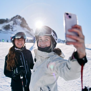 Zwei glückliche Skifahrerinnen auf der Kanzelwand im Kleinwalsertal, eine macht ein Selfie vor winterlicher Bergkulisse bei Sonnenschein. | © Kleinwalsertal Tourismus eGen • Fotograf: Oliver Farys