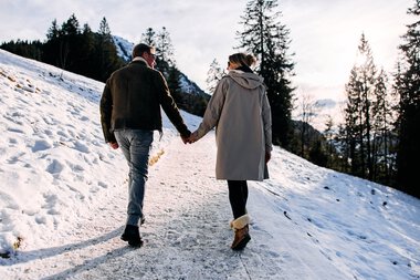 A couple hiking in the snow on a winter walk in Kleinwalsertal, holding hands and enjoying the snowy landscape. | © Kleinwalsertal Tourismus eGen • Photographer: Oliver Farys