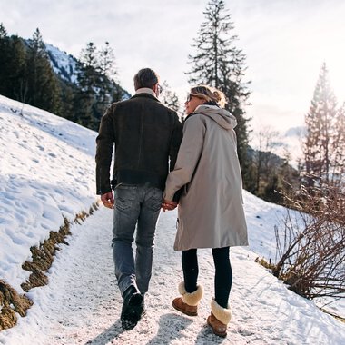 A couple walking in the snow in a winter landscape with trees and mountains in Kleinwalsertal, sunshine. | © Kleinwalsertal Tourismus eGen • Photographer: Oliver Farys