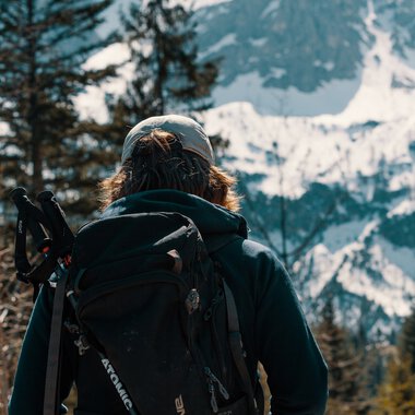 Person with backpack and hiking pole looking at snowy mountains and pine trees in a mountain landscape. | © Kleinwalsertal Tourismus eGen • Photographer: Distillery