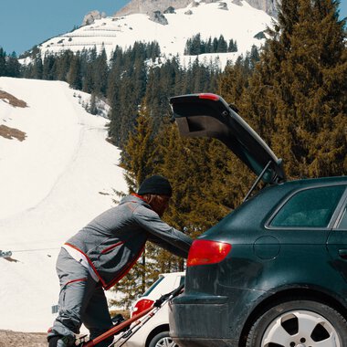 Man preparing skis near a car in a snowy mountain area, with a snow-covered mountain in the background. | © Kleinwalsertal Tourismus eGen • Photographer: Distillery