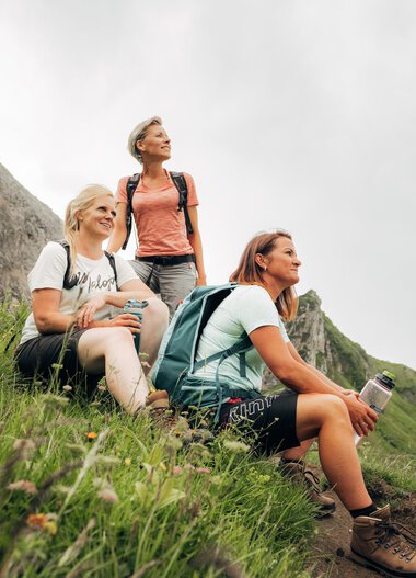 Three women resting during a hike on a green mountain meadow with rocks and flowers, overlooking the surrounding mountains. | © Kleinwalsertal Tourismus eGen • Photographer: Stefan Klauser