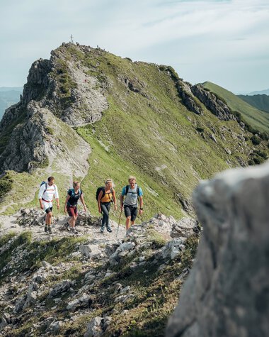 Four hikers on a mountain trail with a view of Kanzelwand in the Alps, surrounded by green slopes and distant mountains. | © Kleinwalsertal Tourismus eGen • Photographer: Stefan Klauser