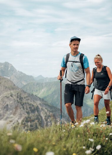 Four hikers walking on a mountain trail with green grass and rugged mountains in the background. | © Kleinwalsertal Tourismus eGen • Photographer: Stefan Klauser