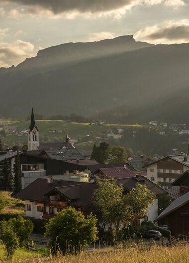 Mountain village with a church and surrounding mountains during sunrise or sunset with warm lighting. | © Kleinwalsertal Tourismus eGen • Photographer: Steffen Berschin 