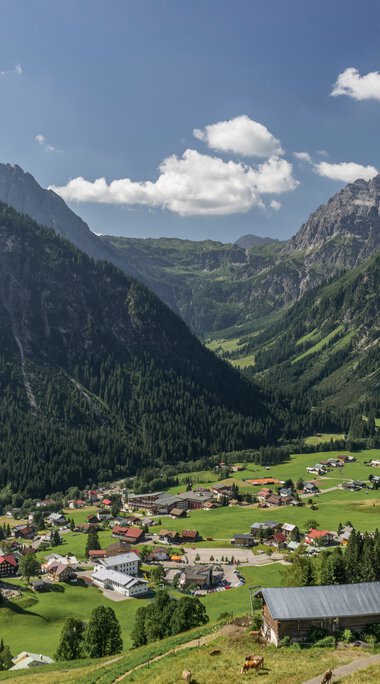 Panoramic view of the village Mittelberg in the Alps, with green hills, mountains in the background, and a blue sky with clouds. | © Kleinwalsertal Tourismus eGen • Photographer: Steffen Berschin 