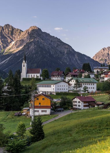 Panoramic view of a small village in the mountains with a church, surrounded by green meadows and trees, against a dramatic mountain backdrop. | © Kleinwalsertal Tourismus eGen • Photographer: Steffen Berschin 