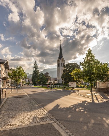 City view with church and sunlight shadows on the street under a cloudy sky in Riezlern. | © Kleinwalsertal Tourismus eGen • Photographer: Stefan Klauser