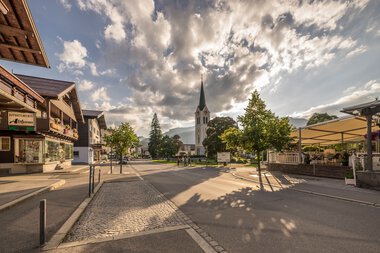City view with church and sunlight shadows on the street under a cloudy sky in Riezlern. | © Kleinwalsertal Tourismus eGen • Photographer: Stefan Klauser