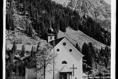 Black and white photo of a small church with a cross, surrounded by trees and mountains, with Wolfgänger in the background. | © Kleinwalsertal Tourismus eGen • Karl Max Kessler Archiv