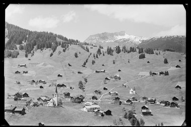 Historical black and white photo of Hirschegg with snow-covered Ifen, surrounded by green fields, scattered houses, and a church. | © Kleinwalsertal Tourismus eGen • Karl Max Kessler Archiv