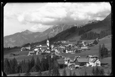 Schwarz-Weiß-Foto des Dorfes Riezlern in den Bergen, mit vielen Häusern, einer Kirche im Zentrum und umliegenden Wiesen und Bergen | © Kleinwalsertal Tourismus eGen • Karl Max Kessler Archiv