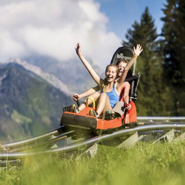 Two girls are laughing and cheering while riding a summer toboggan in the Alps, surrounded by green meadows and mountains. | © Kleinwalsertal Tourismus eGen • Oberstdorf Kleinwalsertal Bergbahnen • Photographer: Bastian Morell