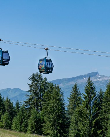 Two cabins of a cable car hang above a green forest landscape with mountains in the background on a sunny day. | © Oberstdorf · Kleinwalsertal Bergbahnen