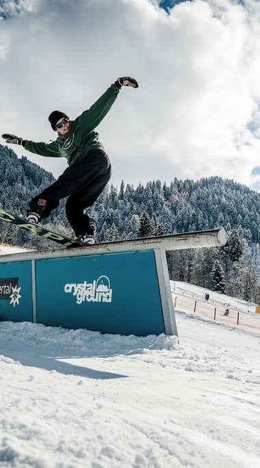Skater beim Rail-Stand auf dem Snowboard in einem Skigebiet mit schneebedeckten Bergen und klarem Himmel. | © Kleinwalsertal Tourismus eGen • Crystal Ground • Fotograf: Stefan Eigner