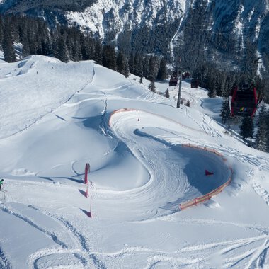 Schneebedektes Wintersportgebiet mit Kurven und Lawinenzaun, umgeben von Wald und Bergen. | © Kleinwalsertal Tourismus eGen • Oberstdorf Kleinwalsertal Bergbahnen