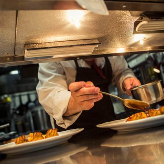 Ein Koch serviert sorgfältig eine Sauce auf vorgerichtete Gerichte in einer professionellen Küche. | © Kleinwalsertal Tourismus eGen • Fotograf: Oliver Farys