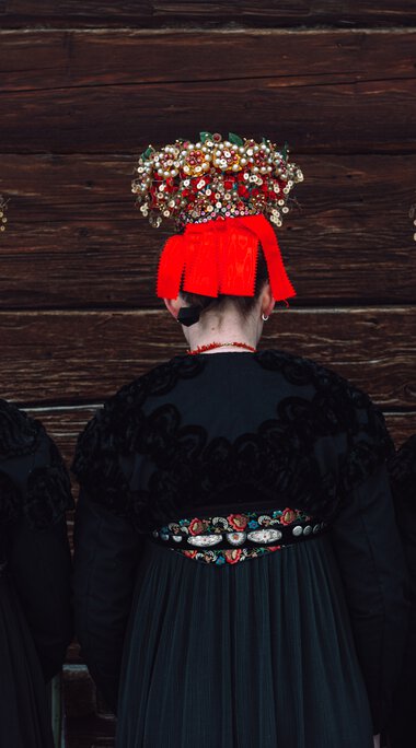 Three women in traditional costumes with colorful headscarves and jewelry standing in front of a wooden wall, back view. | © Kleinwalsertal Tourismus eGen • Photographer: Stefan Klauser