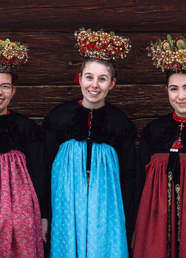 Three young women in traditional costume with elaborate flower crowns on their heads, standing in front of a wooden wall, smiling and wearing traditional attire. | © Kleinwalsertal Tourismus eGen • Photographer: Stefan Klauser