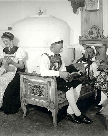 Historical black-and-white photo of five women in traditional costumes, sitting in a living room, engaged in handcrafts and playing music. | © Kleinwalsertal Tourismus eGen