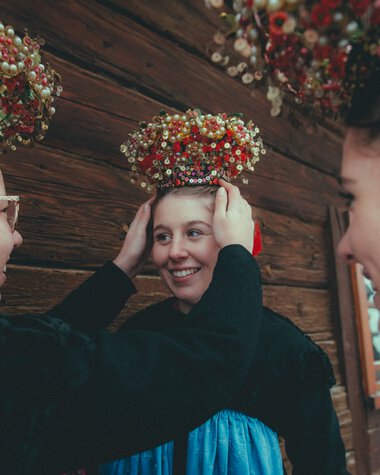 Three women in traditional attire adjusting a festive headpiece on a young woman, with wooden paneling in the background. | ©  Kleinwalsertal Tourismus eGen • Photographer: Stefan Klauser