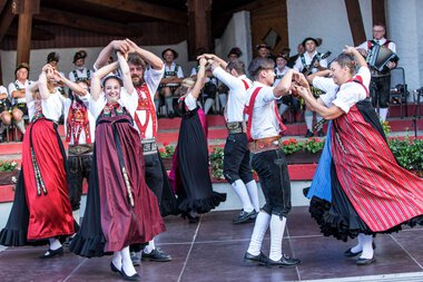 Tanzgruppe in traditionellen Trachten bei einem folkloristischen Fest im Kleinwalsertal, mit Musikern im Hintergrund. | © Kleinwalsertal Tourismus eGen • Fotograf: Frank Drechsel