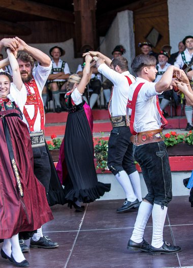 Dance group in traditional costumes performing at a folk festival in Kleinwalsertal, with musicians in the background. | © Kleinwalsertal Tourismus eGen • Photographer: Frank Drechsel