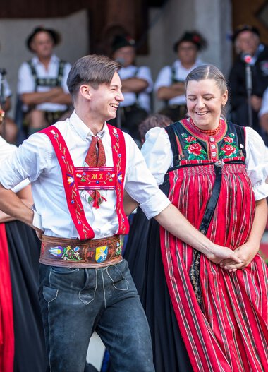 Dancing couple in traditional alpine costumes at an alphorn festival in Kleinwalsertal, surrounded by other dancers in similar attire. | © Kleinwalsertal Tourismus eGen • Photographer: Frank Drechsel