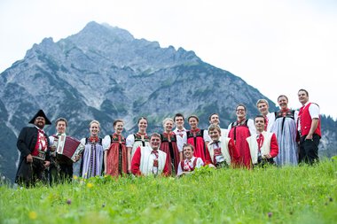 Gruppe von Menschen in traditioneller Kleidung vor einer Bergkulisse, einige mit Musikinstrumenten, im Grünen stehend. | © Kleinwalsertal Tourismus eGen • Fotograf: Frank Drechsel