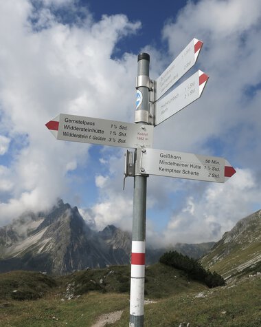 Trail signpost in a mountain landscape with peaks and hills under a cloudy sky. | © Kleinwalsertal Tourismus eGen • Photographer: Antje Pabst