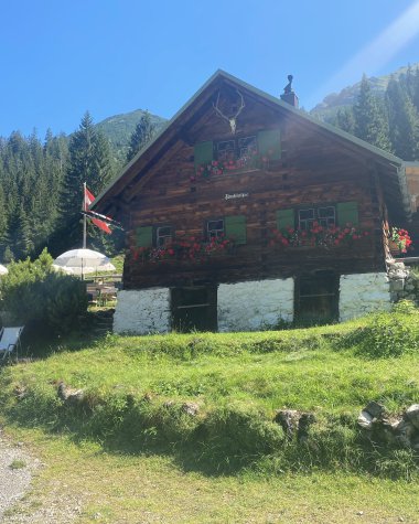 Cozy old wooden chalet with colorful shutters and flowers, in a mountainous landscape on a sunny day. | © Kleinwalsertal Tourismus eGen