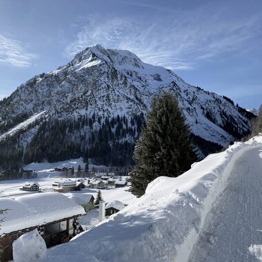 Winter landscape with a snow-covered path, houses, and a large mountain range in the background. | © Kleinwalsertal Tourismus eGen