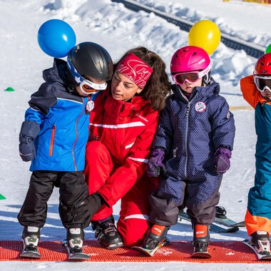 Kinder beim Skischuh-Anziehen im Schnee, mit Skischule-Lehrerin auf einem Wintersportplatz. | © Skischule Bödmen Baad