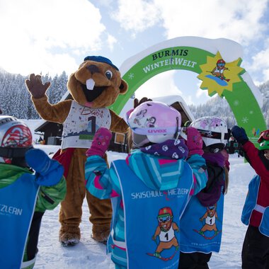 Kinder beim Skikurs in Riezlern begrüßen den Burmi-Winterwelten-Maskottchen vor einem Winterhintergrund mit Bergen. | © Kleinwalsertal Tourismus eGen • Photographer: Oliver Farys