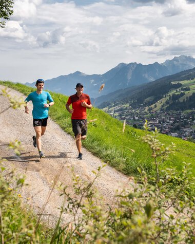 Two runners trail running on a narrow path in the mountains, with a green landscape and mountain view in the background. | © Kleinwalsertal Tourismus eGen • Fotograf: Rupert Mühlbacher