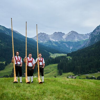 Three men in traditional costumes standing in a green meadow with alphorns, mountains, and a forested valley in the background. | © Kleinwalsertal Tourismus eGen • Photographer: Oliver Farys