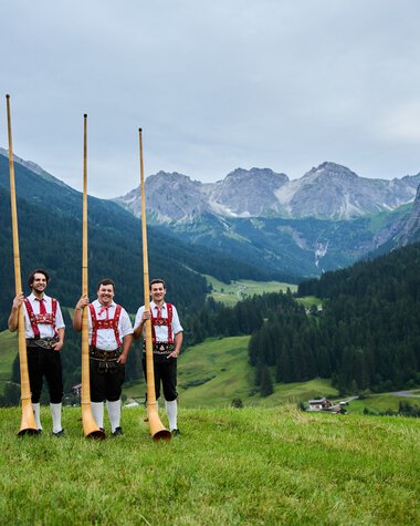 Three men in traditional costumes standing in a green meadow with alphorns, mountains, and a forested valley in the background. | © Kleinwalsertal Tourismus eGen • Photographer: Oliver Farys