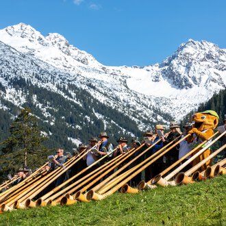 Group photo at the 2024 Alphorn Festival with musicians in traditional clothing and alphorns, against a stunning mountain backdrop with snow-capped peaks. | © Kleinwalsertal Tourismus eGen • Photographer: Frank Drechsel