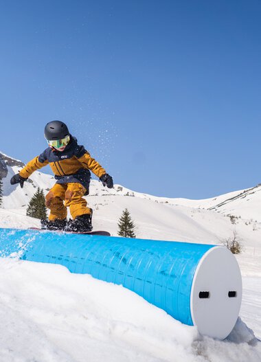 Ein Kind beim Snowboarden im verschneiten Gebirge mit blauer Himmel, das auf einem gebogenen, blauen Rail fährt. | © The Grilos • Chris Gollhofer